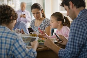 A mother, a father and two children are looking at their cell phones and tablets while sitting together at the dinner table.