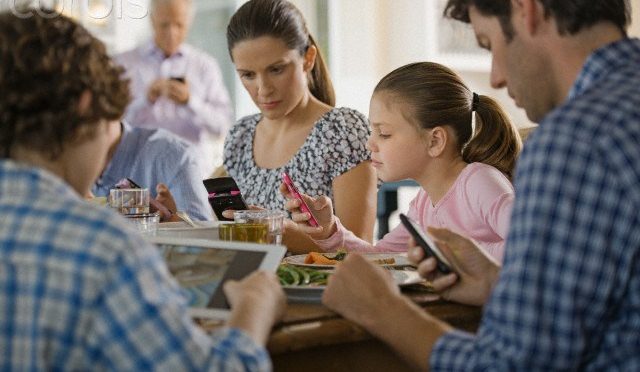 A mother, a father and two children are looking at their cell phones and tablets while sitting together at the dinner table.