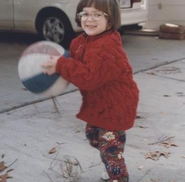 This is an image of Sara Luterman as a child. She is wearing a red sweater and leggings. She has brown hair and glasses. Sara is holding a basketball that is larger than her head.