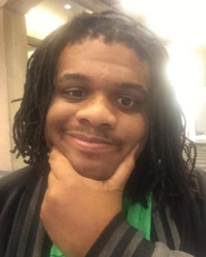 This is an image of Timotheus Gordon. He is a black man with medium length dreadlocks and a light mustache. He is smiling and holding his chin in his hand. 