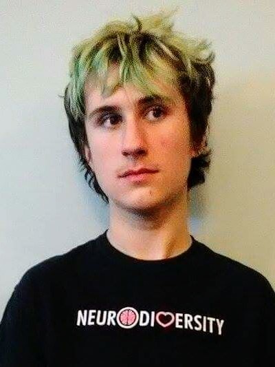 This is an image of Niko Boskovic, from the shoulders up. He is a white teenage boy with brown and blue hair. He is wearing a t-shirt that says "neurodiversity" on it and looking off to the side.