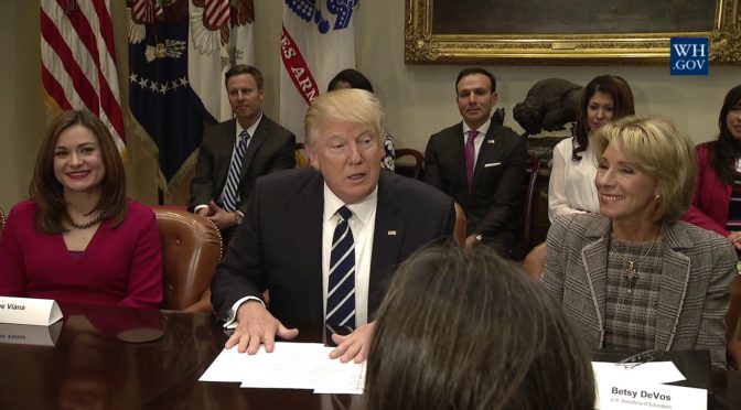 This is an image of Donald Trump sitting at a table and speaking Betsy DeVos is sitting to his right.