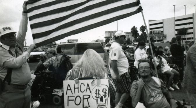 This is a black and white image of ADAPT of Texas protesting in Austin. There is an American flag with the stars in the shape of an accessibility symbol in the background. There is also a crowd of people. One member of the crowd is holding up a sign that reads "AHCA FOR" and then is followed by a skull with dollar signs for eyes.