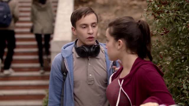 This is an image of Sam, the main character of Atypical. He is a white teenage boy wearing black headphones around his neck. He is talking to a white teenager girl.
