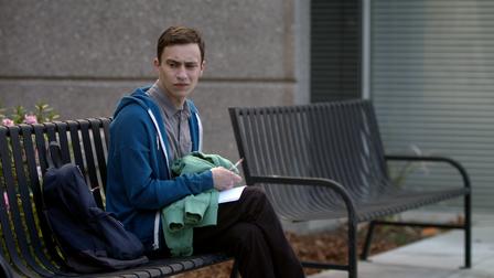This is an image of Sam, the main character of Atypical, sitting on a public bench. The bench is metal. Sam is a white teenage boy with dark hair. he is wearing a hoodie.