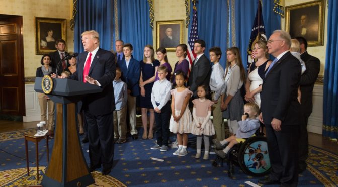 This is an image of President Trump speaking at a podium. Behind him, "Obamacare Victims" stand and listen, except for one small boy who is in a wheelchair. Secretary of Health and Human Services Tom Price stands at the ight edge of the photograph.
