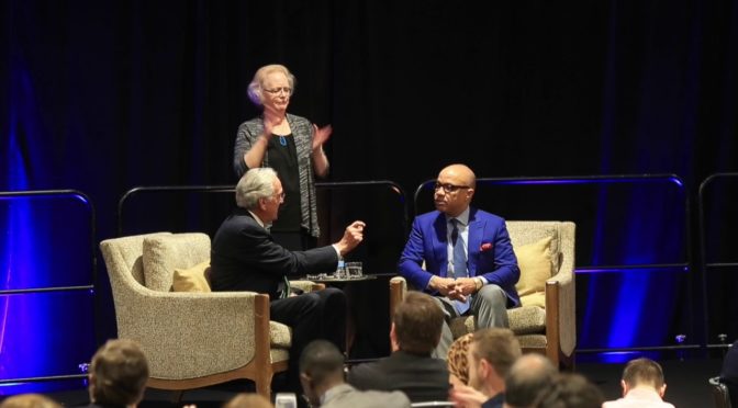 This is an image of Ford Foundation President Darren Walker and former Senator Tom Harkin sitting on a conference stage. A sign language interpreter stands between them. Darren Walker is a slim, bald black man with glasses. He looks like he is listening intently to Senator Harkin. Senator Harkin is an older white man with gray hair. Both men are wearing suits and ties.